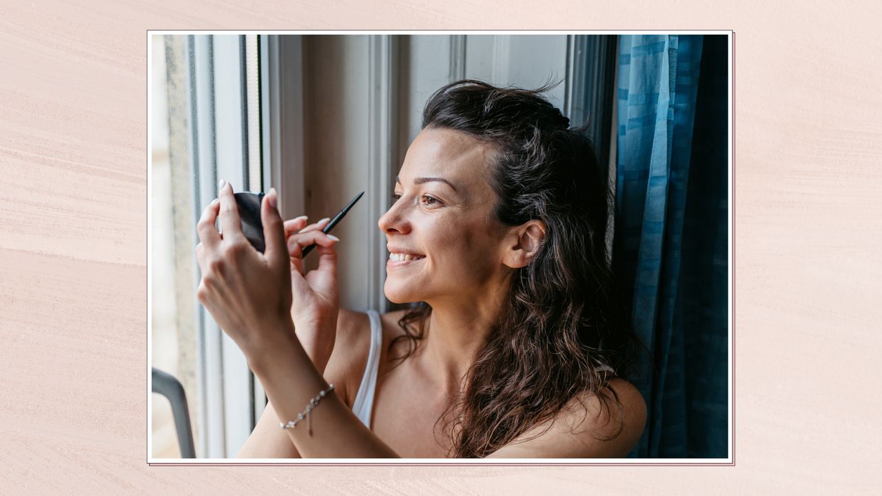 Side view of woman sat in front of a window applying eyeliner pencil while looking into a compact mirror 