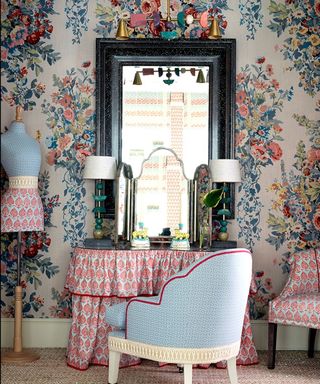 Colorful dressing room with floral wallpaper, wooden dressing table with patterned fabric skirt, upholstered chair, mirror, table lamps