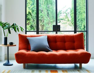 Habitat’s Kota three-seater sofa bed – a large orange/red three-seater sofa with a high back, on a white floor in front of black crittall doors, with a wooden and black side table next to it with a black mug and plant on it