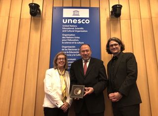 three people hold a small metal disk inside a frame while posing for a picture in front of a sign that reads "unesco"