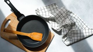 A frying pan and wooden spatula on a wooden chopping board, beside a tea towel