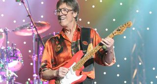 Hank Marvin of the Shadows plays his red strat and wears and orange and black graphic shirt.