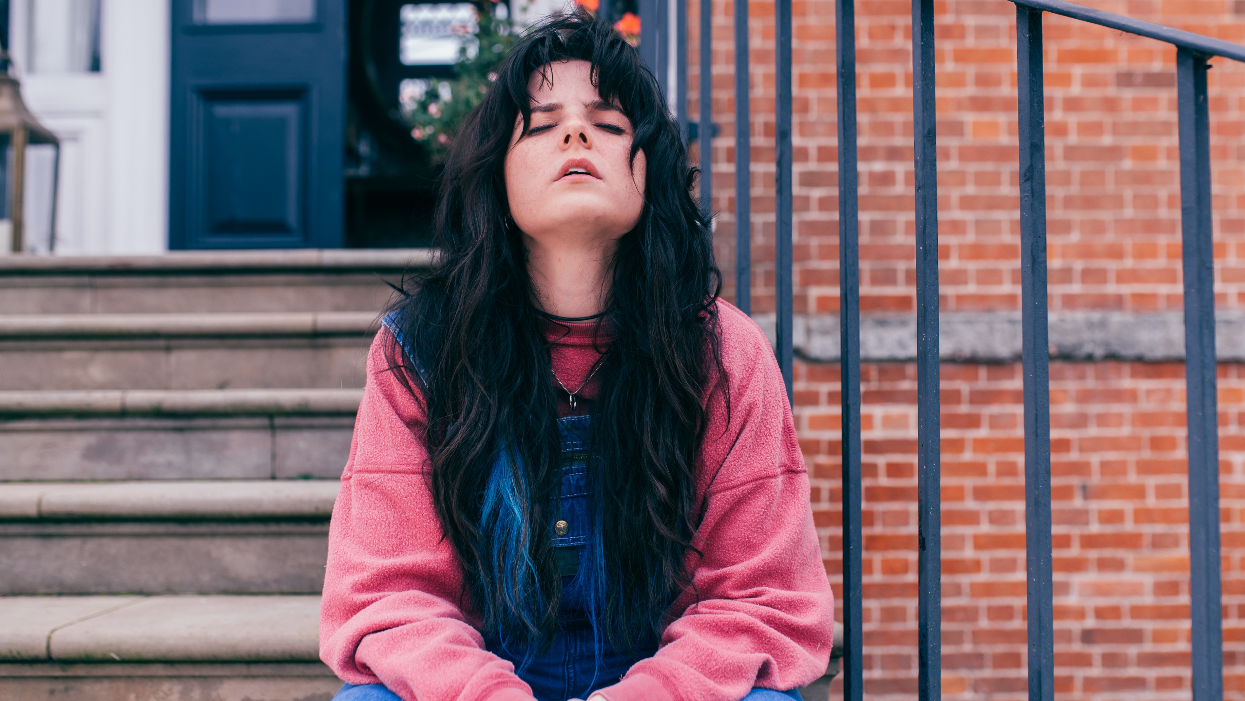 Ria (GABRIELLE CREEVY) looking stressed sitting on some steps leading up to a house.