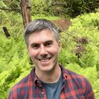 Headshot: James Holland in front of a bushes and a tree, wearing a red and blue patterned shirt and a blue t-shirt