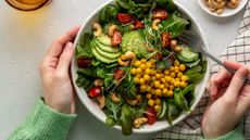 Bowl of leafy vegetables, chickpeas and fibre-rich foods with woman's hands around the bowl and tea towel on the counter to represent the process of making postbiotics