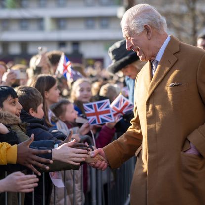 King Charles wearing a camel colored coat shaking hands with excited children outside who are waving Union Jack flags