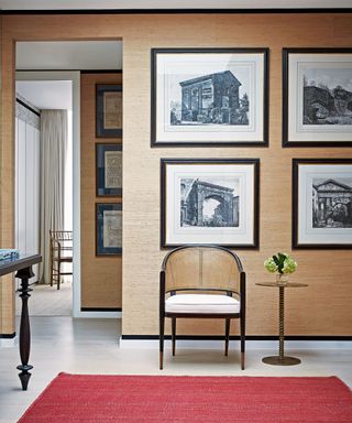 Hallway with skylight, tiled floor and red rug, framed photographs above a chair and occasional table.