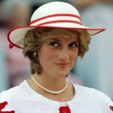 A headshot of Princess Diana smirking wearing a white hat with red trim and pearls