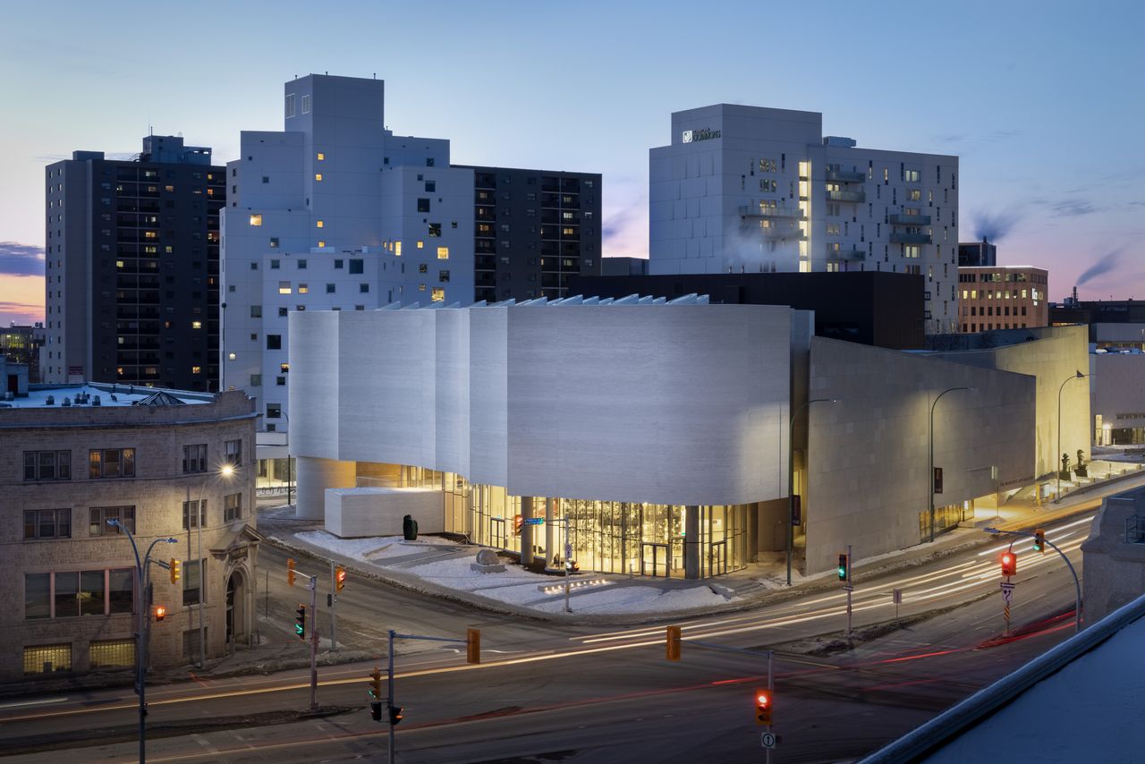 View of the exterior of the quamajuq inuit art centre, set beyond a road with traffic lights, tall buildings lit up in the distance, small chimneys with steam on some of the roof tops, clear dusk sky