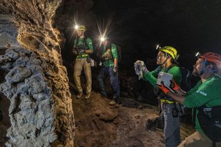 People in green shirts and lit hardhats point devices at a rock formation.