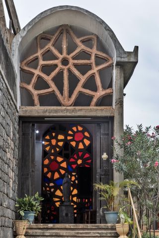 Daytime exterior image of the Dominican chapel in Ibadan features many carefully handcrafted elements, including the 12 stained-glass flowers behind the statue of the Virgin Mary and 12 carved columns, potted plants and flowered shrubs, stone steps, pale blue cloudy sky