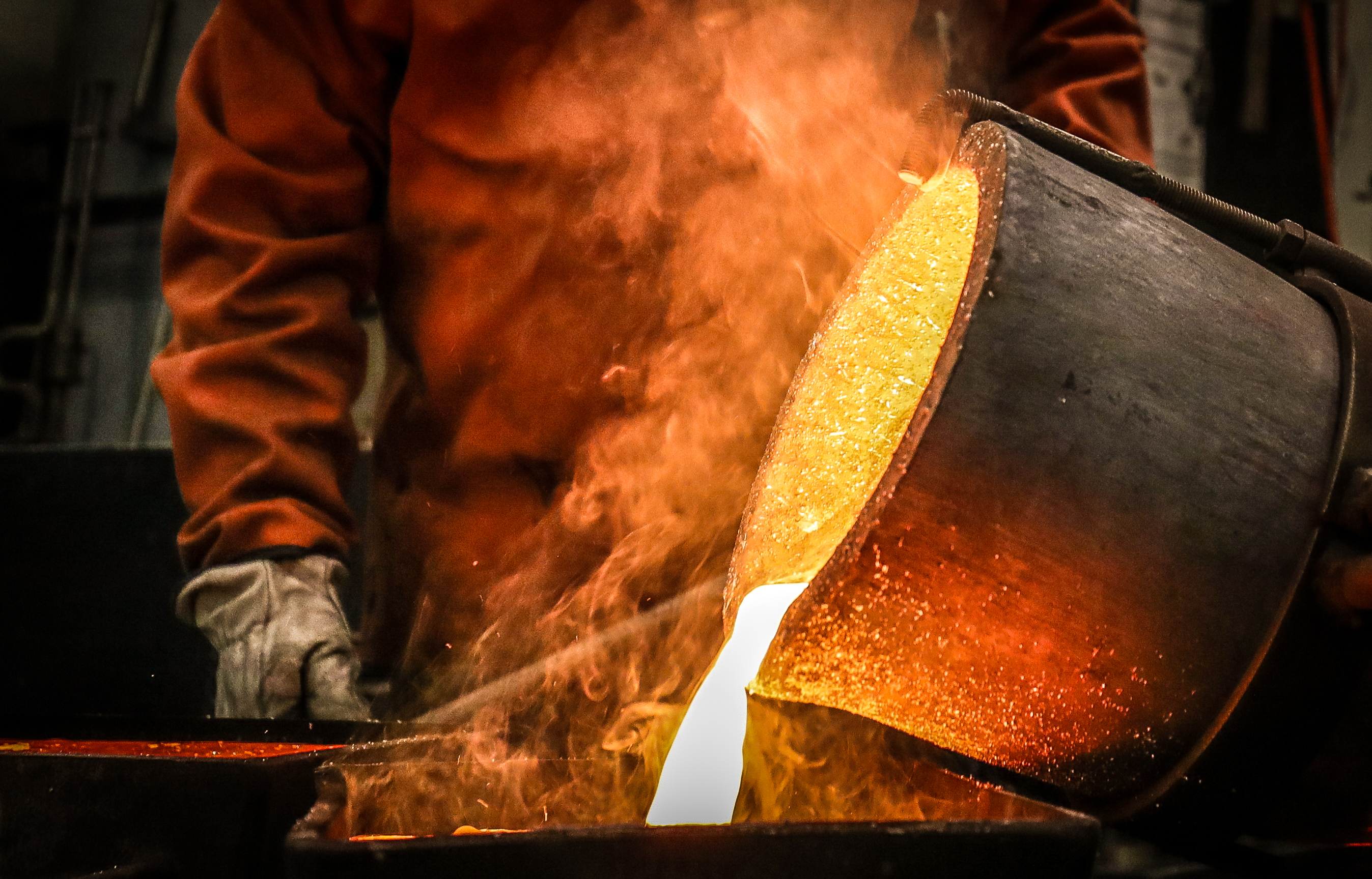 Close-up shot of molten gold being poured by a large pot onto a table. A man, holding the pot, is wearing a heavy-duty apron and gloves