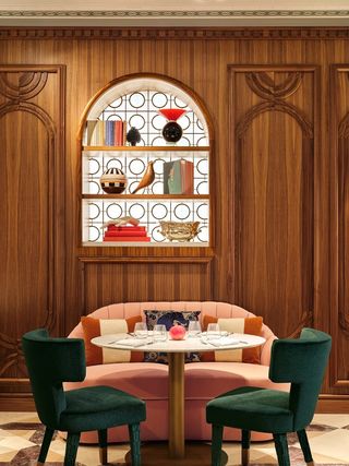 A plush, retro-futuristic hotel restaurant features a lip-shaped pink velvet banquette and two green velvety chairs positioned around a round marble table, wood-drenched walls with a decorative, arched shelving unit filled with collectibles, and terrazzo flooring.