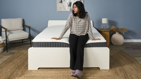 Mattress tester Becky George sits on the middle of the Emma Hybrid Airgrid mattress on a bed frame in our sleep studio, testing the edge support and comfort of the mattress