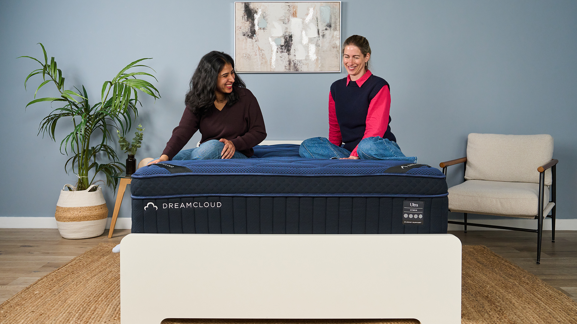 The image shows two mattress testers sitting on top of a navy blue mattress, placed on a white bed frame in a muted blue bedroom.