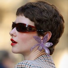 A woman at fashion week with a pixie haircut wearing sunglasses, large floral earrings and red lipstick