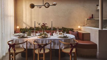 A table setting with candles and lots of foliage as well as tableware such a utensils, plates and napkins. By the dinner table are dining seats with red ribbons wrapped around it 