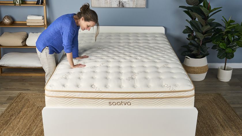 A woman stands next to the Saatva Classic Mattress on a bed frame in a bedroom, pressing her hands into the medium-firm surface to test the pressure relief