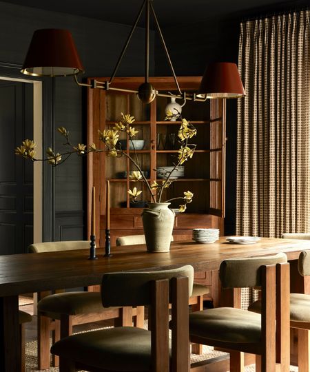 A dramatic dining room with dark charcoal walls and a long wooden table. A brass chandelier with red shades hangs above a rustic ceramic vase with yellow blossoms, with a tall wooden china cabinet in the background.