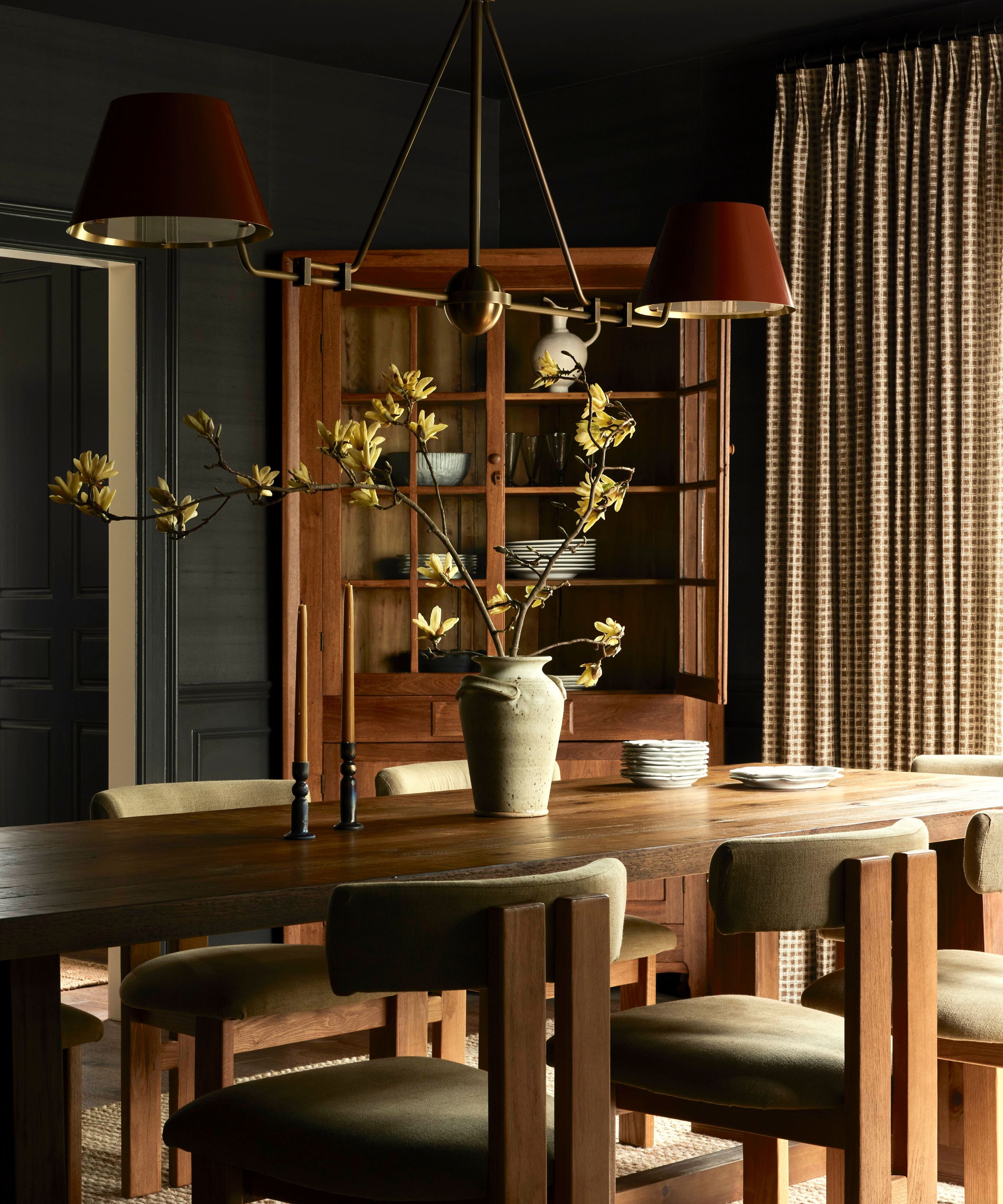 A dramatic dining room with dark charcoal walls and a long wooden table. A brass chandelier with red shades hangs above a rustic ceramic vase with yellow blossoms, with a tall wooden china cabinet in the background.