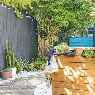 Stepping stone pathway surrounded by large gravel with trees and planters on either side and festoon lighting on the fence