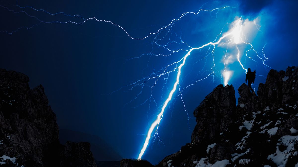 A person hikes on a mountain as a lightning strike hits overhead against a dark blue sky.