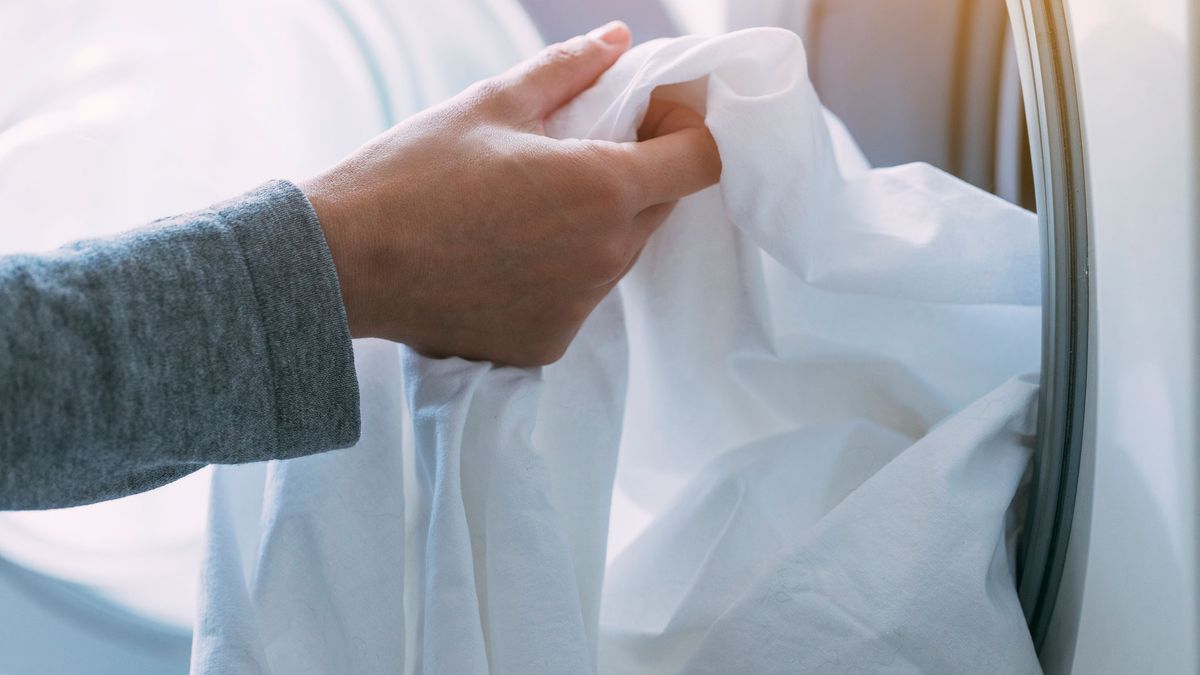 How often should you clean your sheets: Image shows a hand putting white sheets into a front load washer.