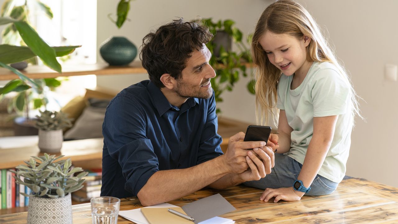 A dad and his young daughter look at his smartphone together at the kitchen table.