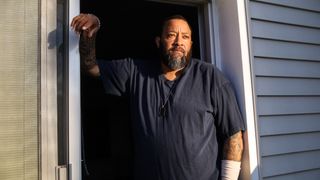 Kidney transplant recipient Corey Mayes, a middle-age Black man with a beard and wearing a black t shirt, stands in the doorway of his home.