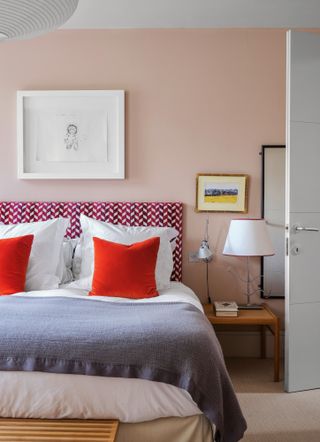 Bedroom with light pink walls, red-pink patterned headboard, white bedding and bright red cushions