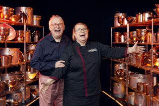 Christopher Biggins and his mentor Rosemary Shrager stand between two racks of cooking equipment, both grinning wildly