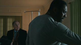 Idris Elba looks out the window as Dermot Crowley stands holding his coat in Luther: The Fallen Sun.