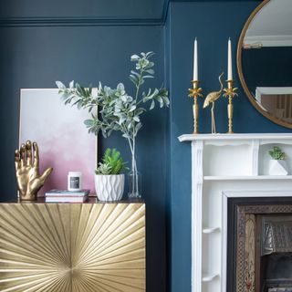 Living room with dark blue walls, honey oak floor, brown patterned fireplace with white mantel, gold cabinet, and decorative touches in gold, pink, white and green