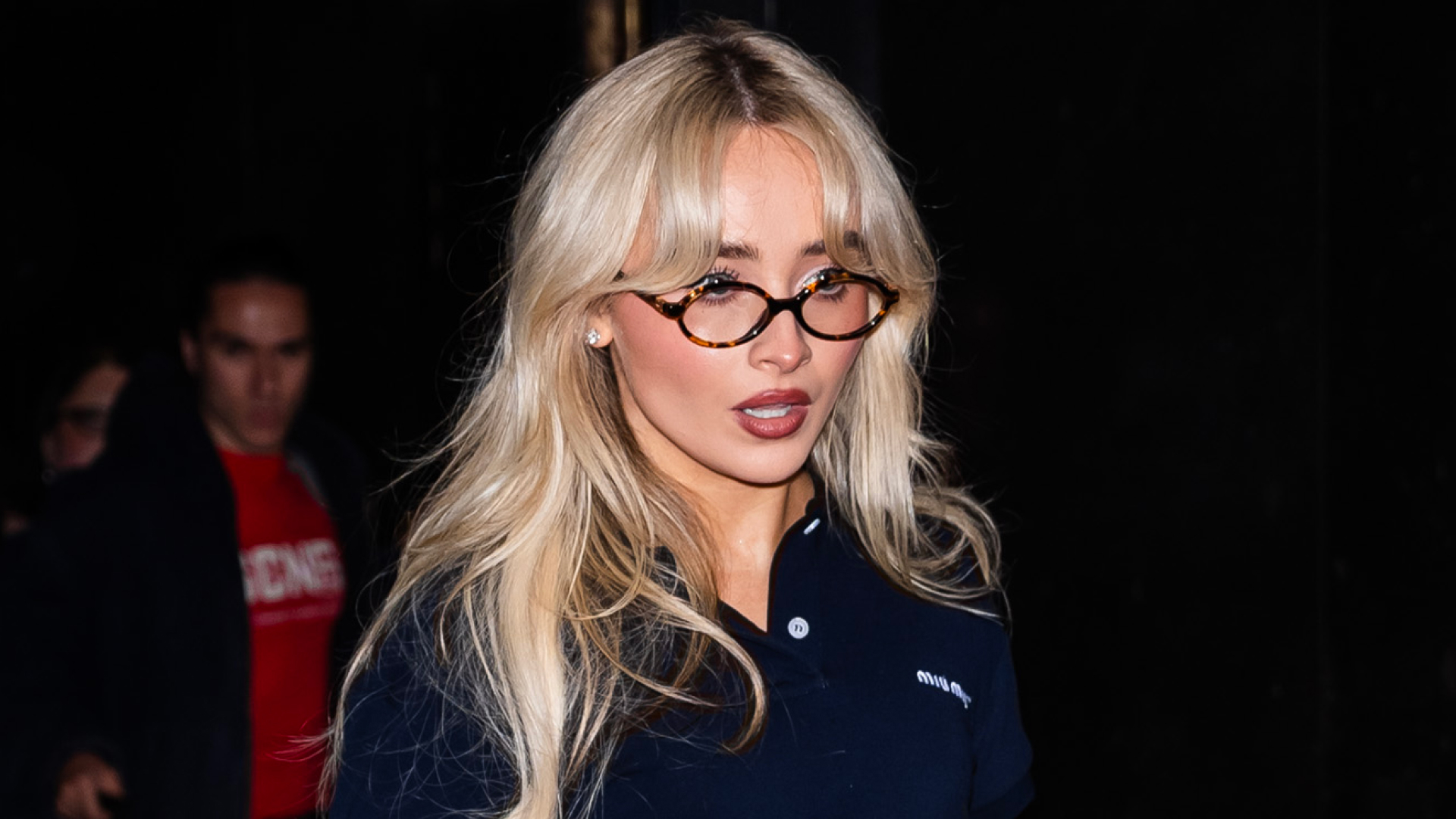 Sabrina Carpenter wearing a black polo shirt with tortoiseshell glasses and long blonde hair