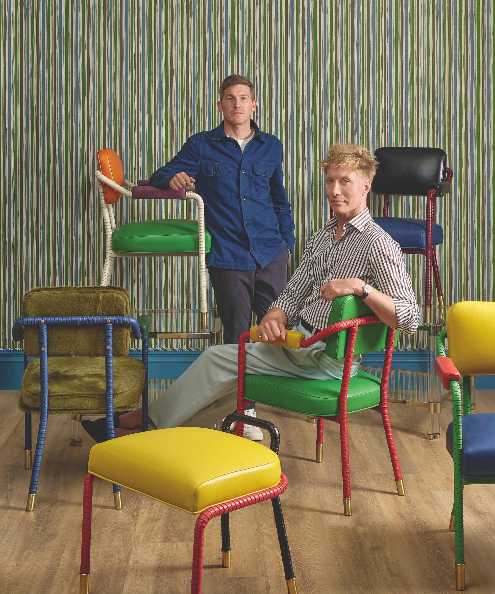 A collection of different coloured chairs. One man sits on a green chair and behind another man stands beside a chair
