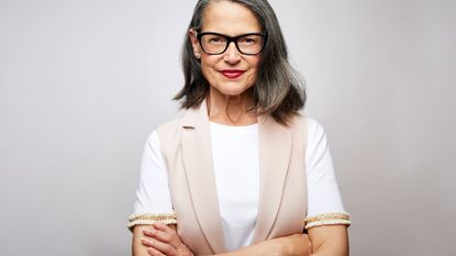 An older woman stands with a small smile and folded arms.