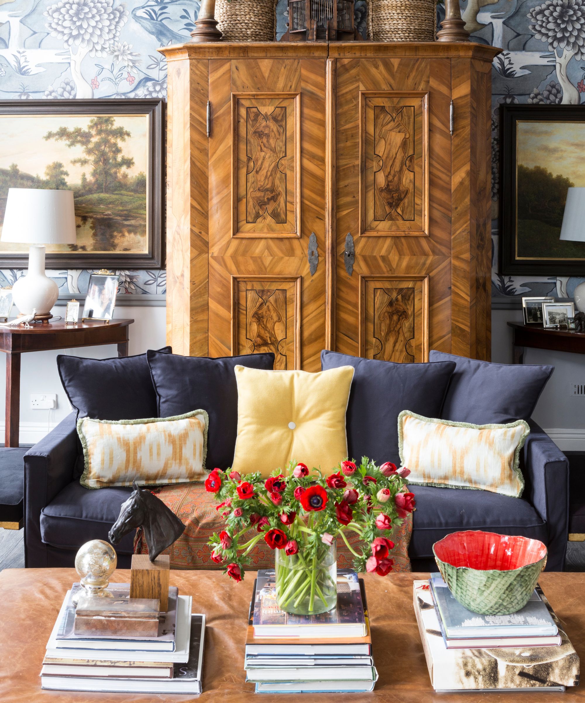 Living room with large burl wood cabinet, a blue sofa, yellow pillows, and a marble coffee table with stacks of books, a vase of roses, and a red and green bowl.