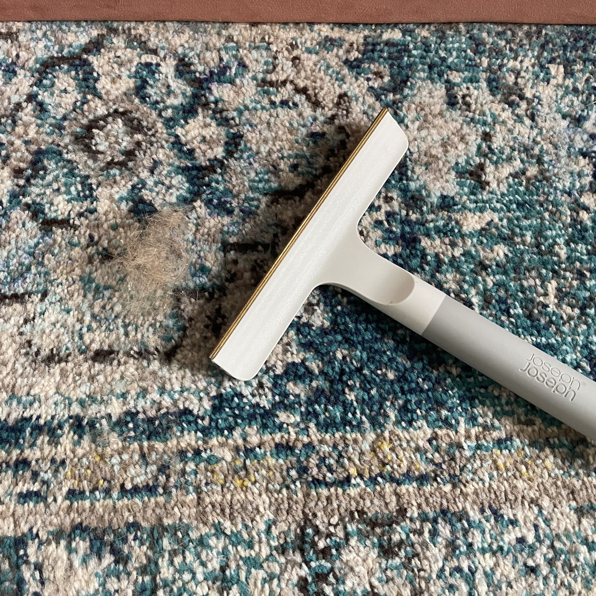Joseph Joseph pet hair carpet scraper tool on a blue rug, with a pile of hair next to it