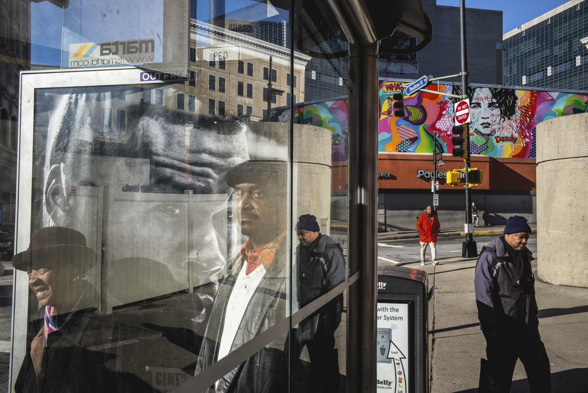 A busy street corner seen through the glass of a bus shelter, its reflections layering a smiling woman in a brown hat, a man in a flat cap and a giant black-and-white advertisement face over a mural