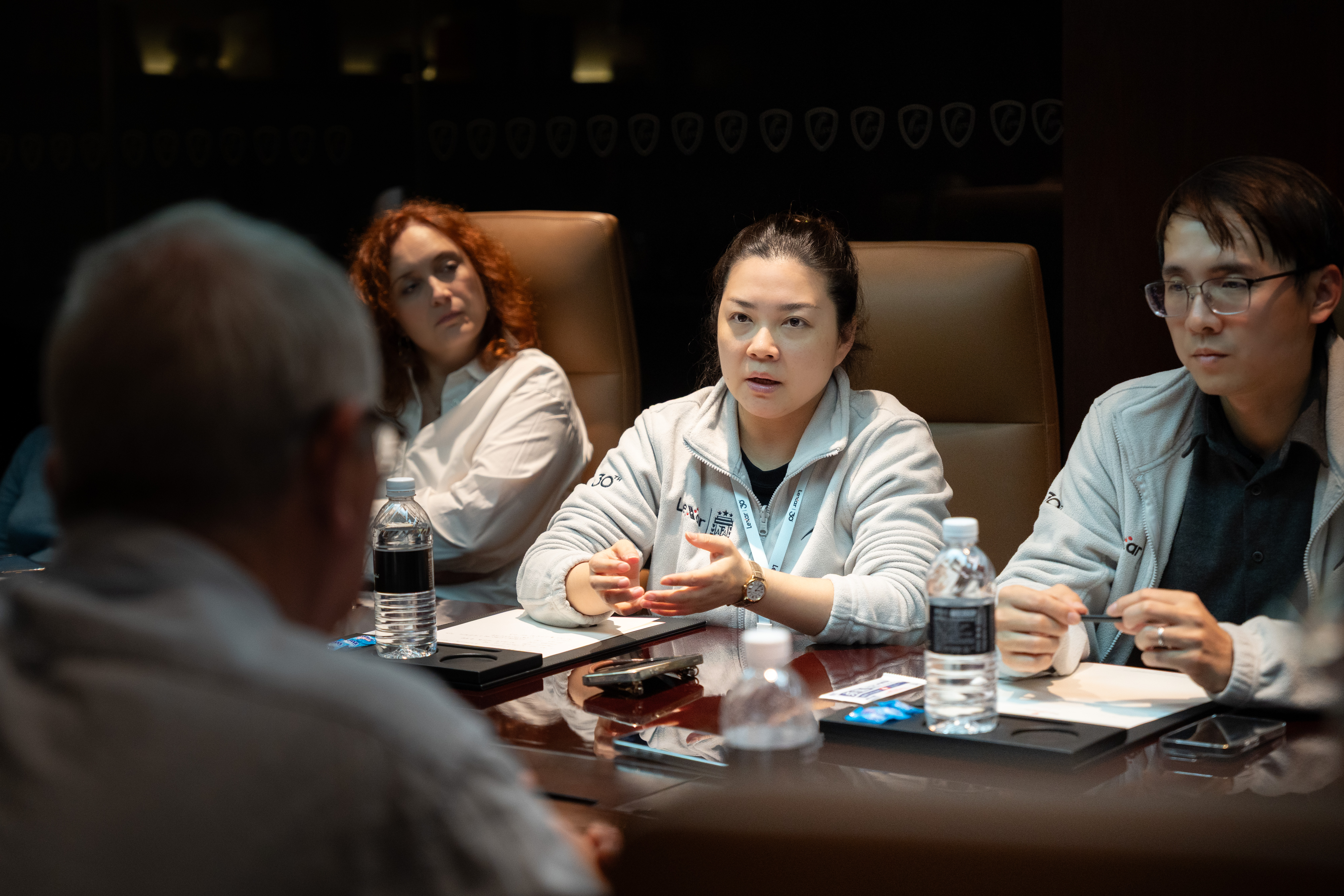 A woman seated at a dark conference table gestures while speaking to colleagues during a press conference