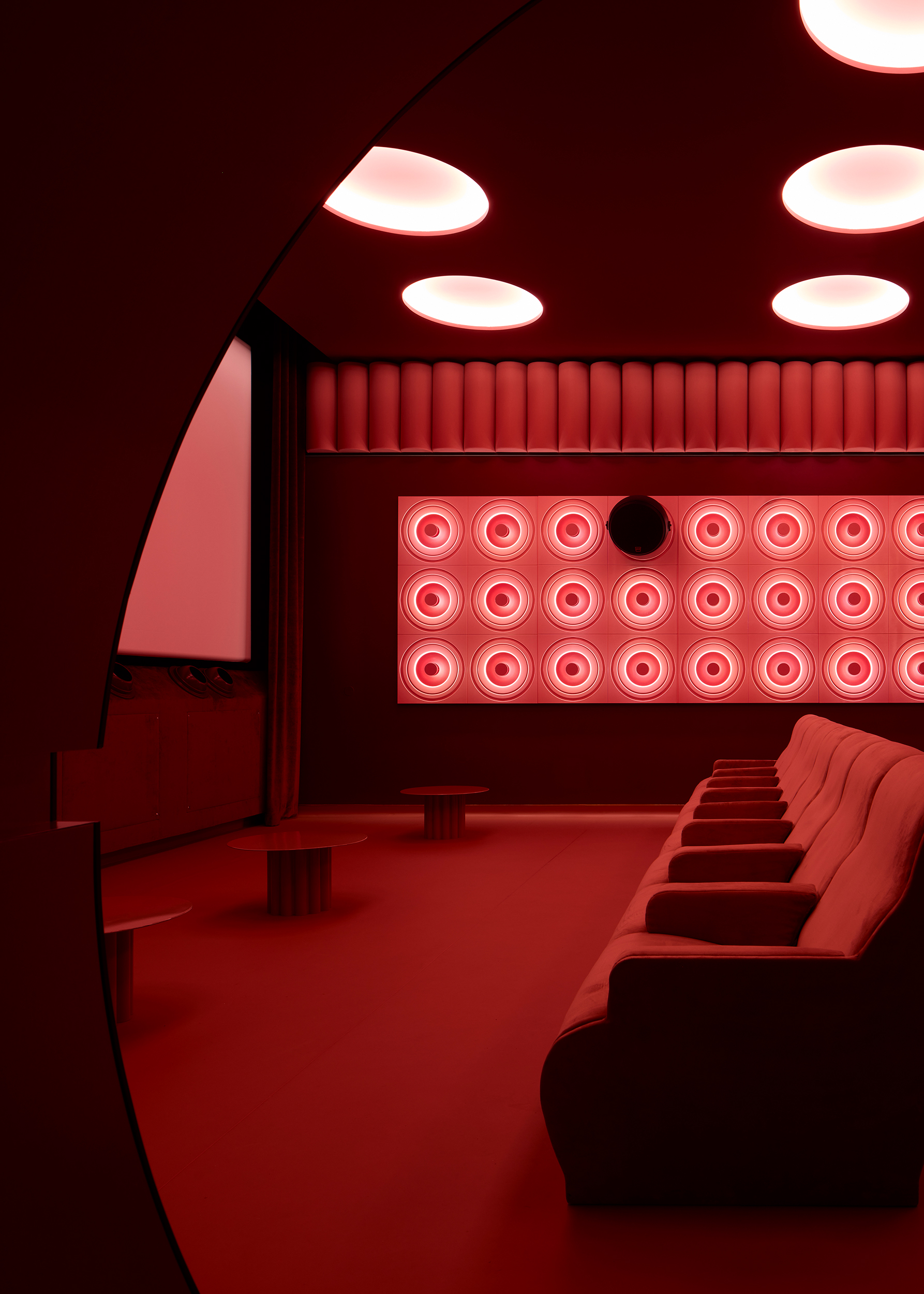 A cinema screening room with velvety red seats, porthole window-like ceiling lights, high-tech sound panels in pink, rounded shapes, and semi-circular portals.