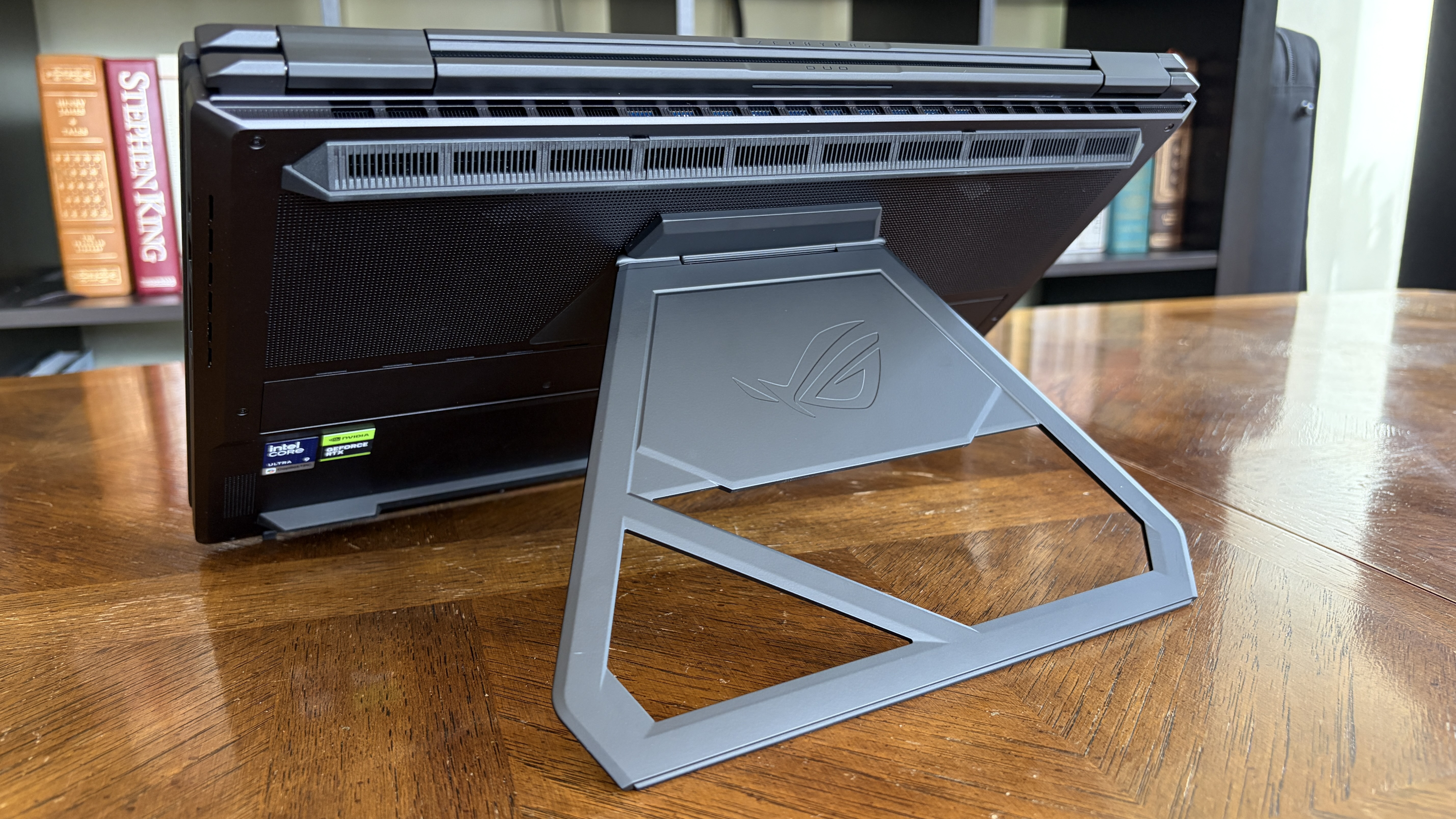 A gaming laptop is propped open on a stand, showing a sleek design with a back cover and set on a wooden table. Books are neatly arranged in the background.