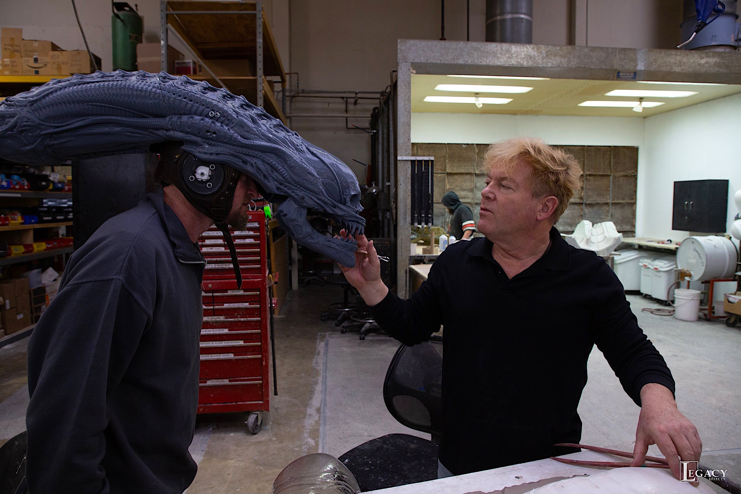 a performer wears an alien monster headpiece while another man inspects it 