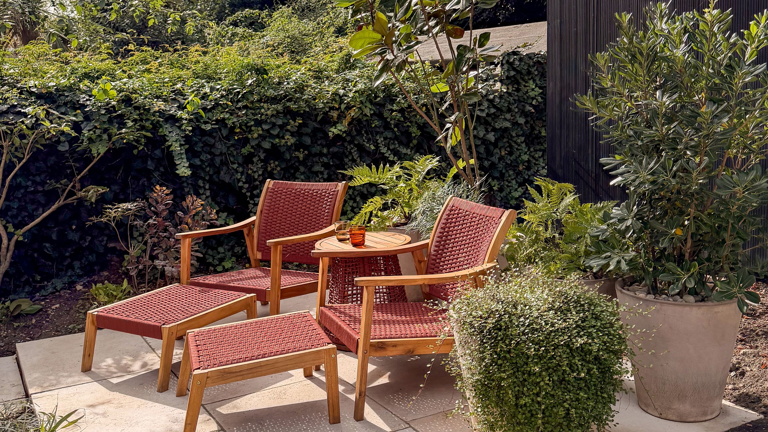 Two wooden and read woven recliner chairs with some table on patio area by hedge in garden