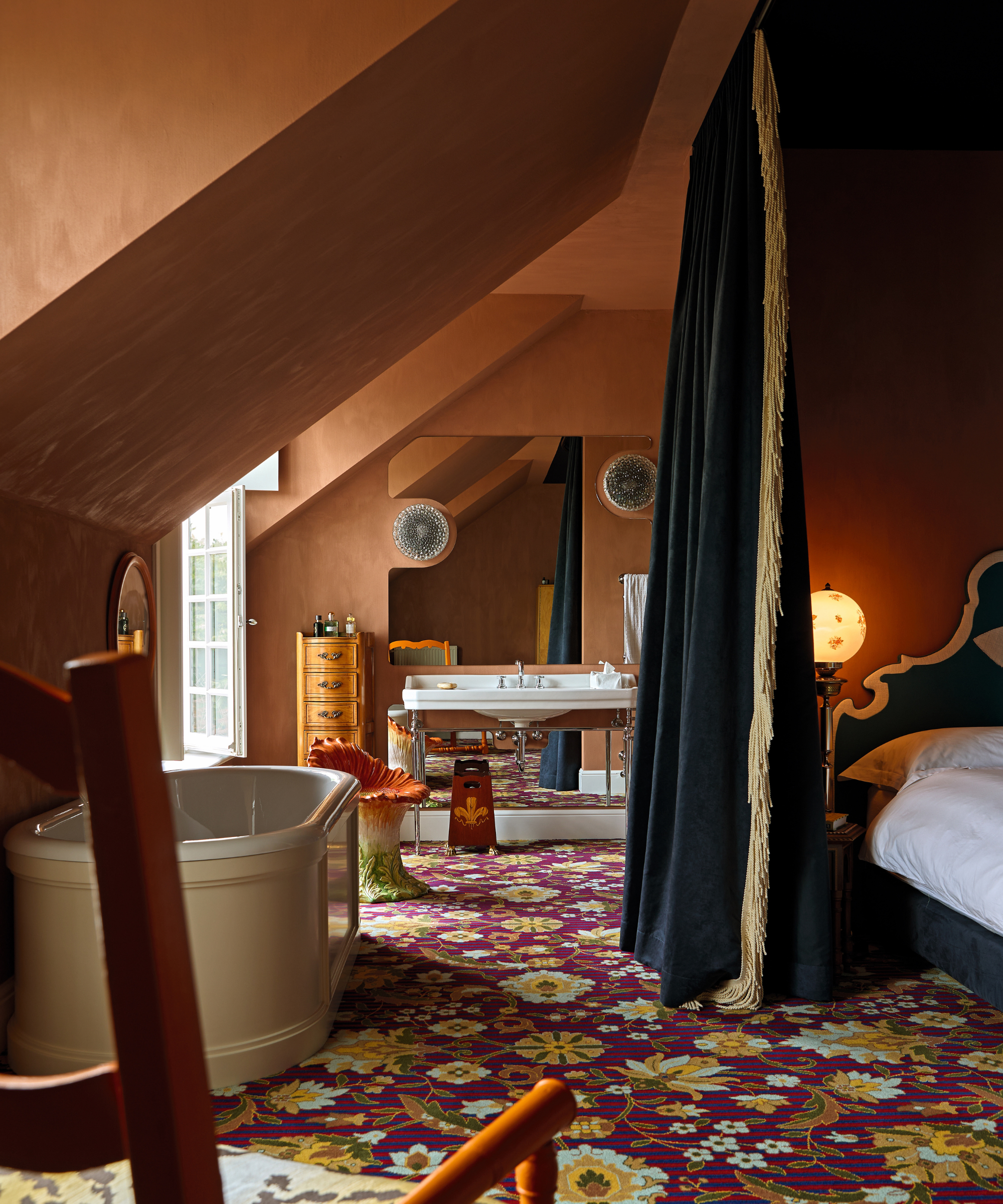Bedroom in attic with orange walls and bed with navy velvet curtain, patterned floral carpet and bath in room