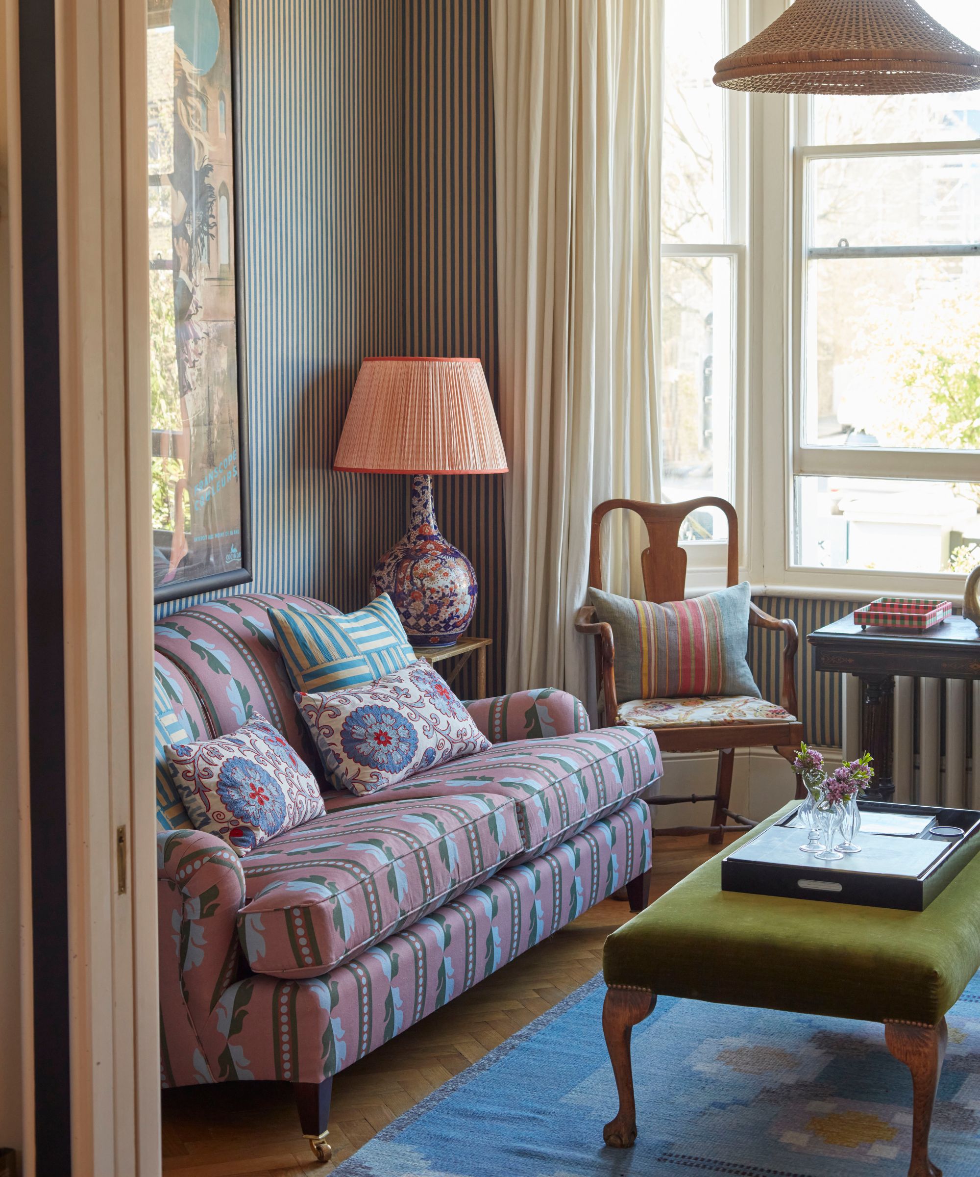 a striped small living room with a large bay window with a pink floral upholstered sofa, blue vintage rug and a green velvet ottoman