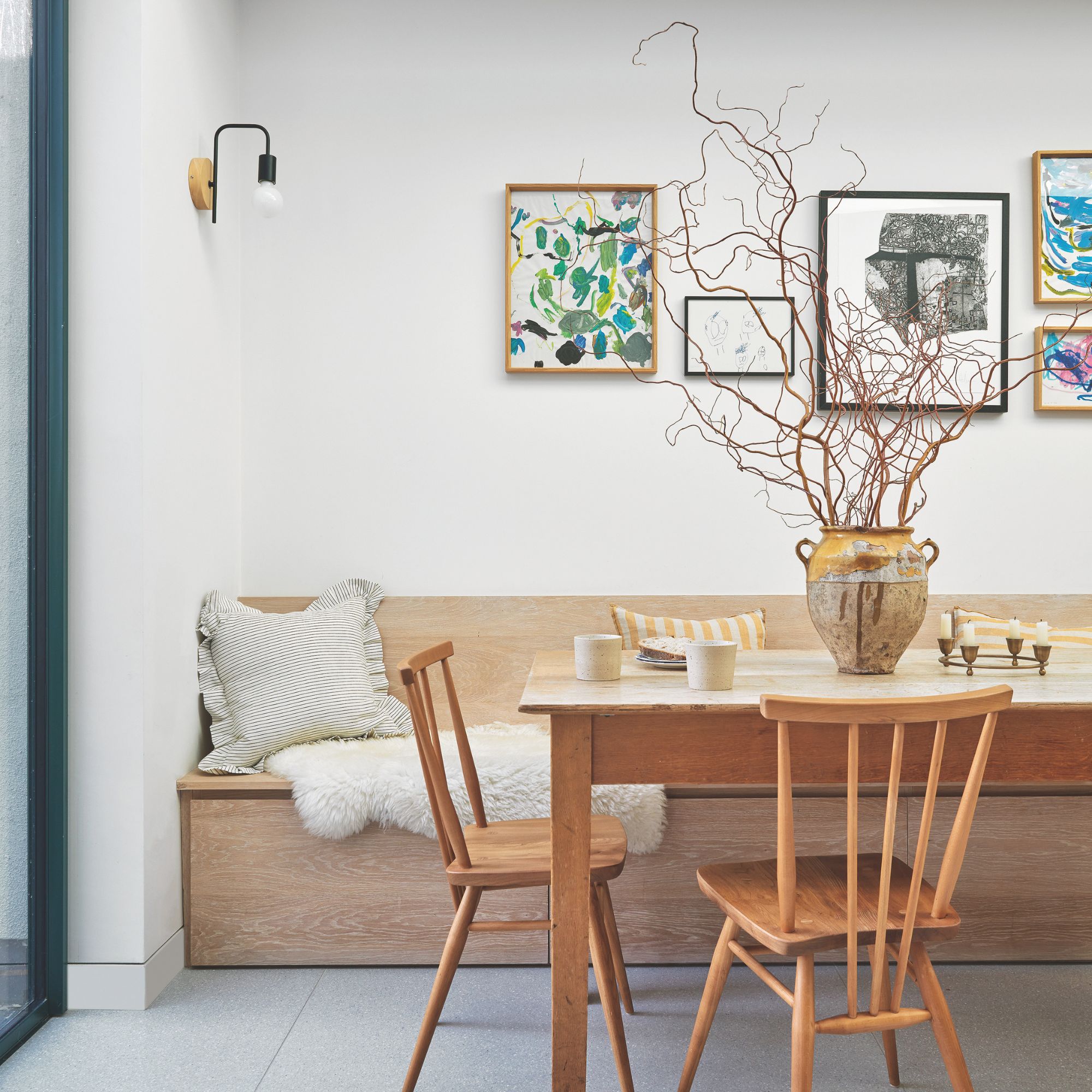 White painted dining room with wooden dining table and a banquette bench behind it. There's a vase of decorative twigs on the table