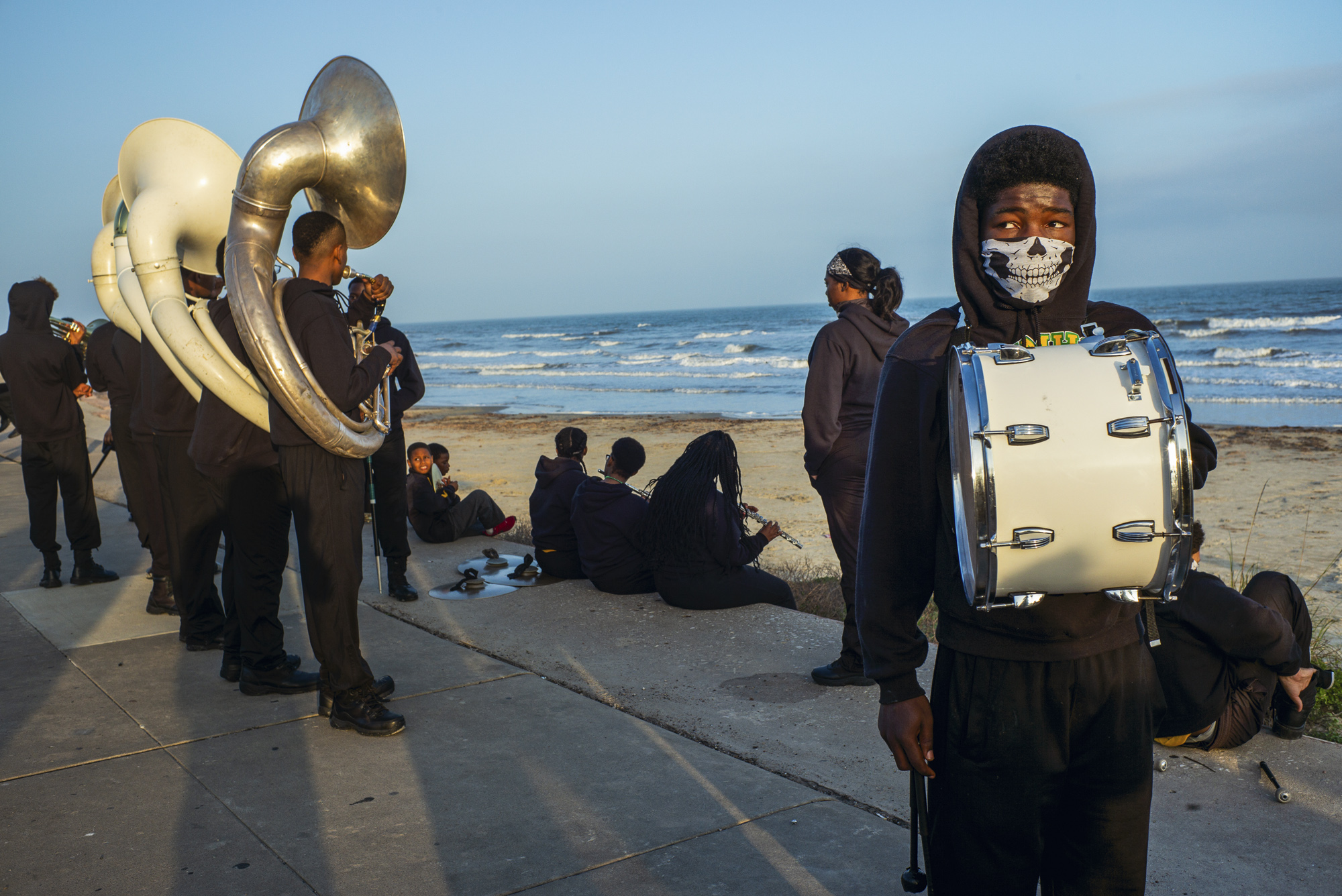 Members of a marching band in black hoodies wait on a seafront promenade at golden hour, with sousaphone players on the left, others sitting on the sea wall beyond and a drummer in the foreground