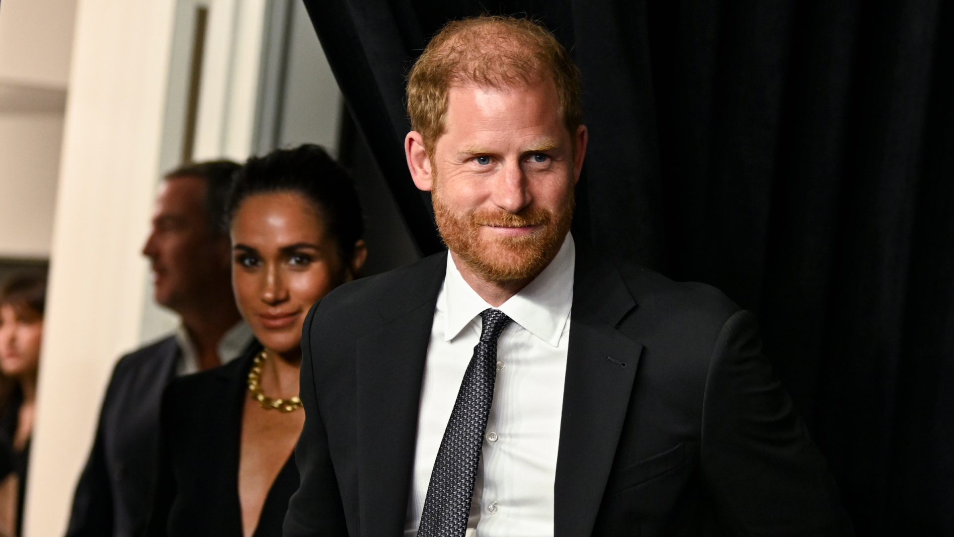 Prince Harry wears a black suit and has red hair and is holding hands with wife Meghan Markle who is wearing a black shirt dress