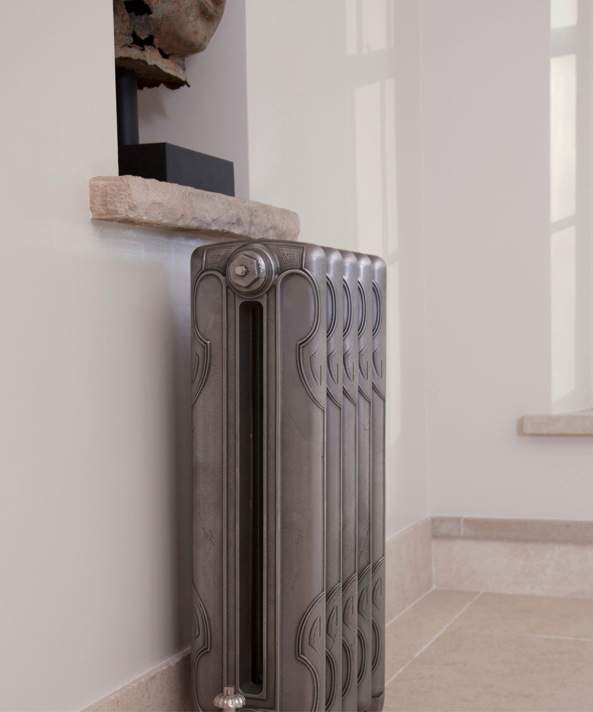 An ornate cast iron radiator in a brushed grey finish, adding a touch of period character to a neutral hallway with stone flooring.